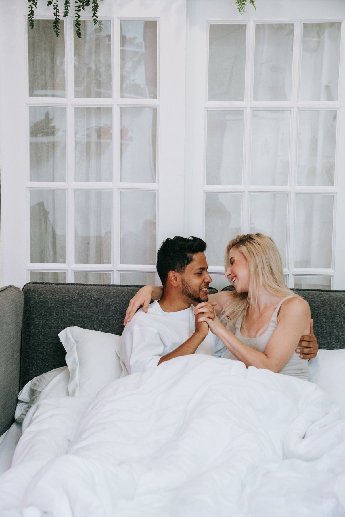 Two adults sitting together under white bedding on a gray sofa bed.