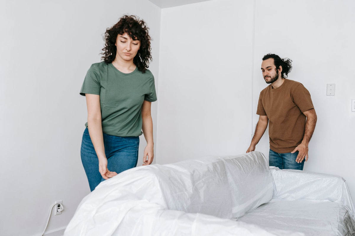 Two people spreading a white covering over the back and arms of a sofa.