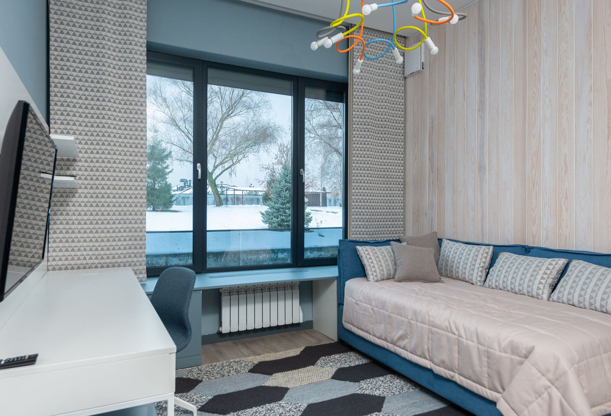 Blue upholstered daybed-style sleeping area with a white quilted coverlet and loose pillows beside a window and desk.