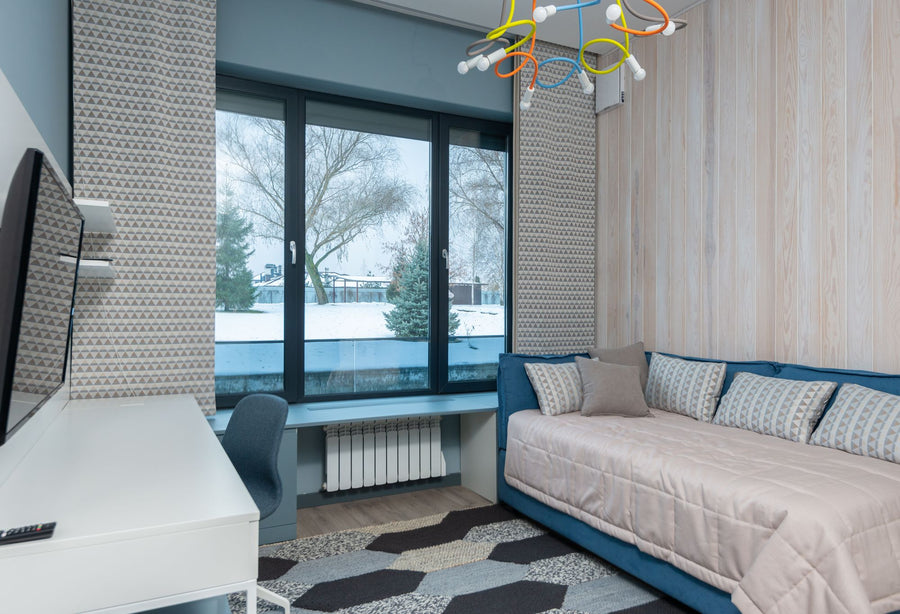 Blue upholstered daybed-style sleeping area with a white quilted coverlet and loose pillows beside a window and desk.