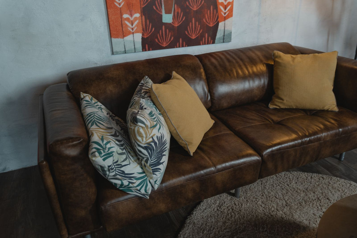 Dark brown leather sofa with mustard and pale leaf-patterned pillows against a light blue-gray wall.