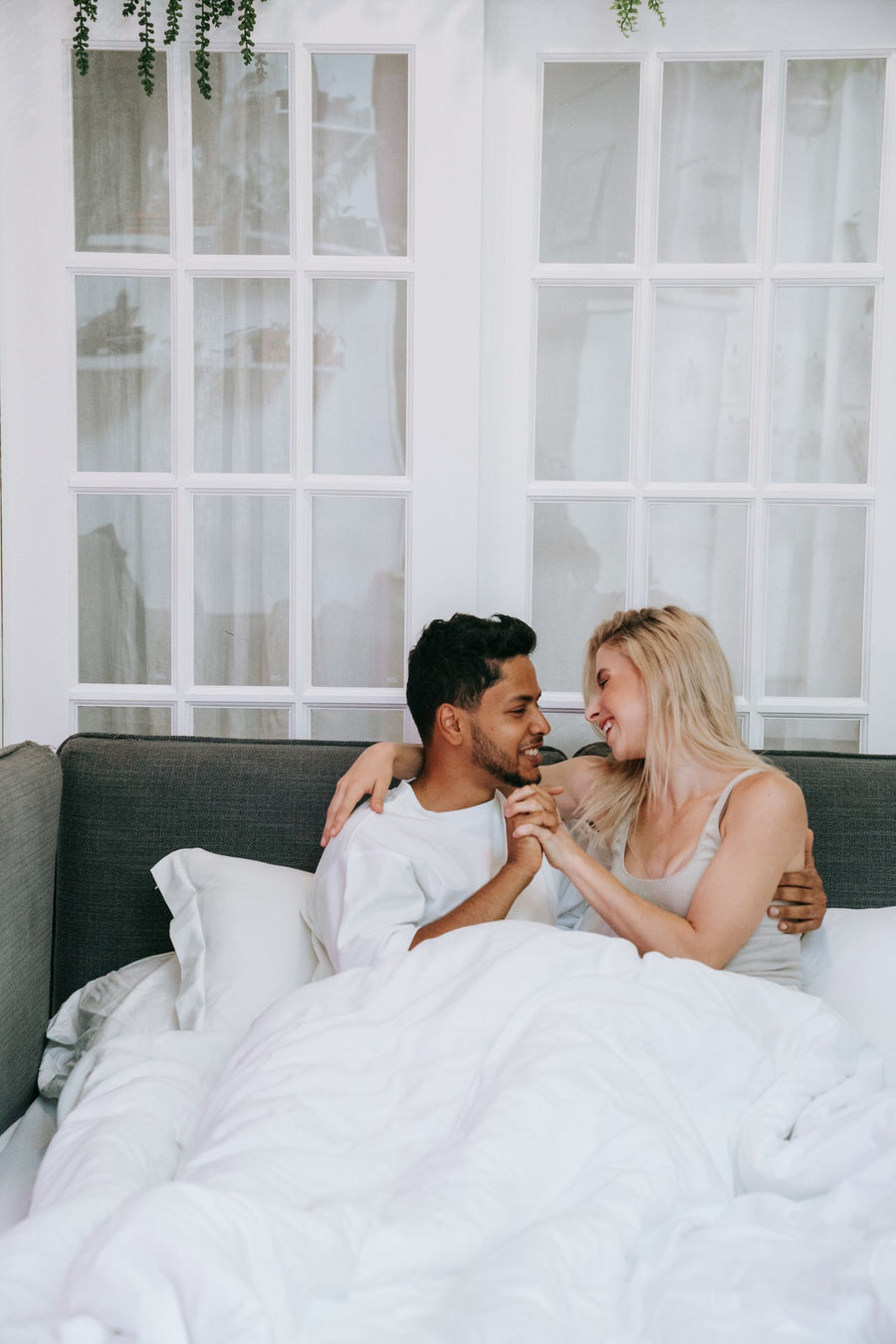Two adults sitting together under white bedding on a gray sofa bed.
