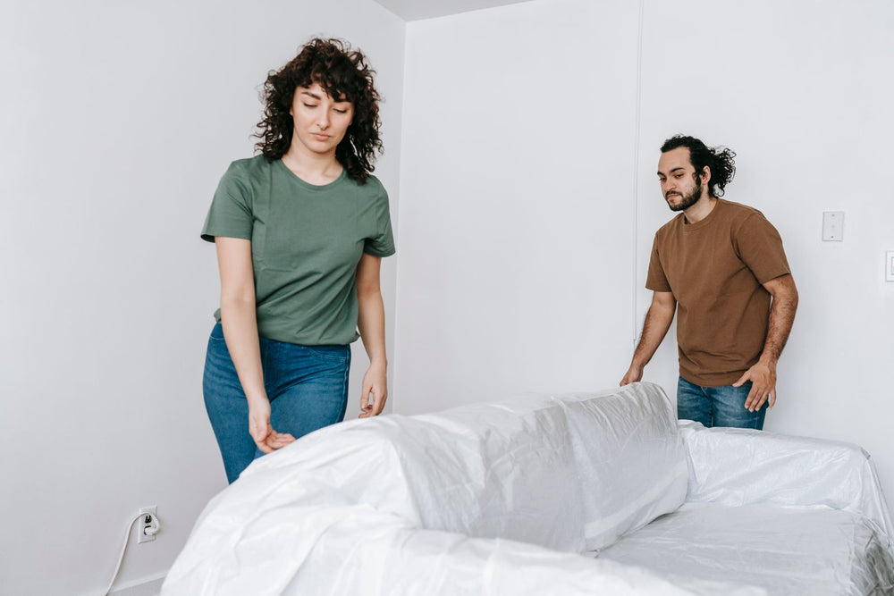 Two people spreading a white covering over the back and arms of a sofa.