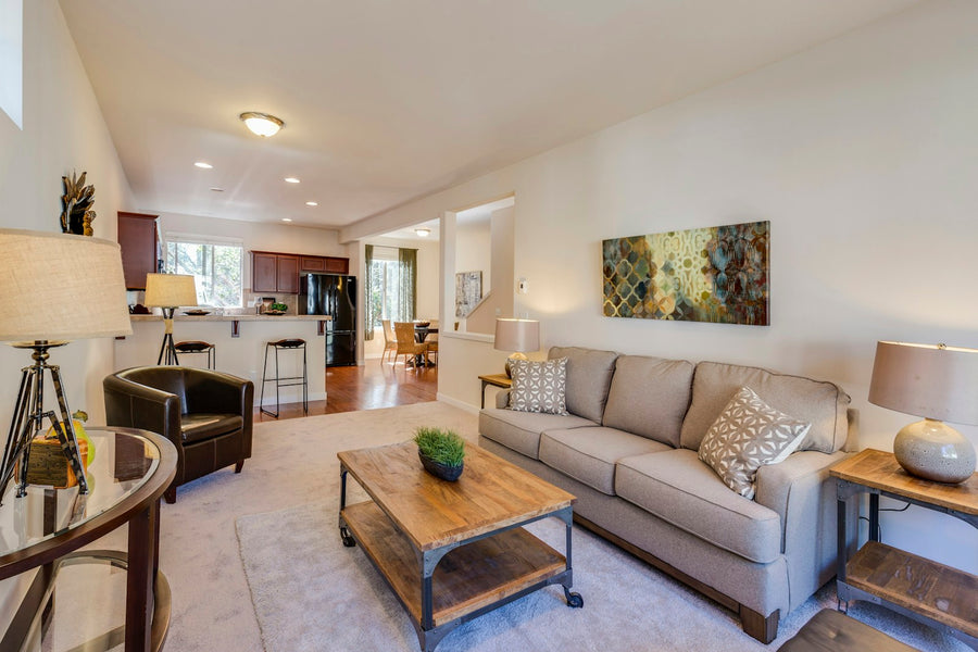 A gray sofa, separate dark armchair and wood tables in a living room opening toward a kitchen