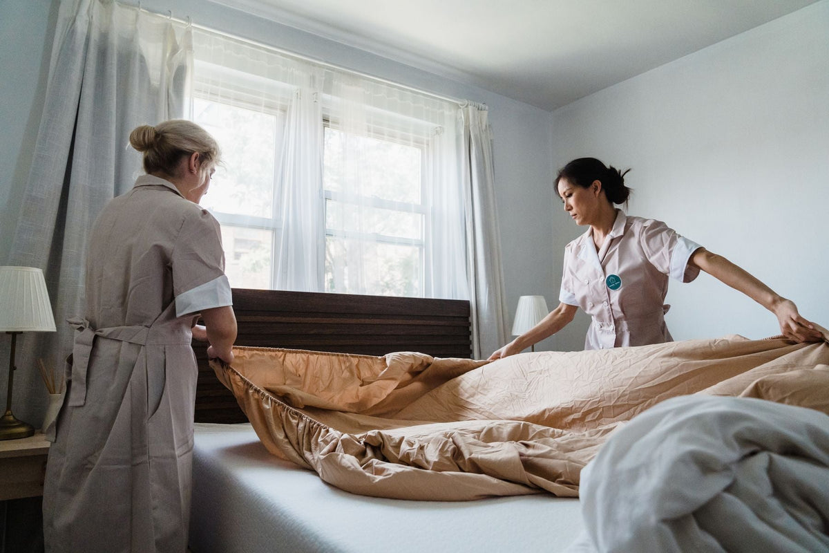 Two people spreading a tan fitted sheet with a gathered elastic edge over a white mattress.