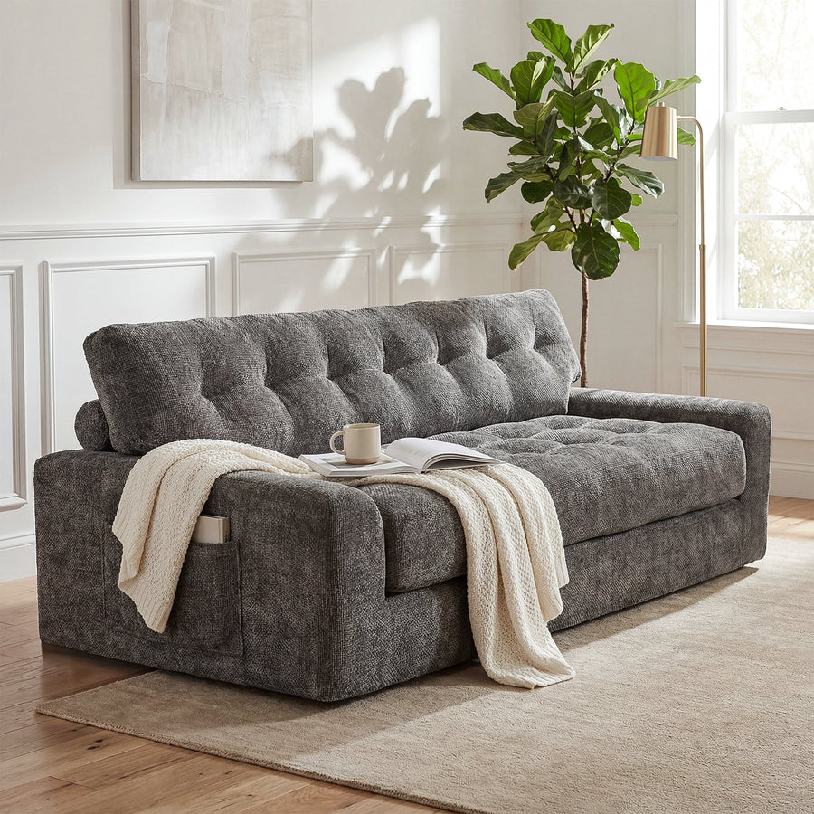 Dark gray upholstered sleeper sofa shown closed in a bright room with a throw, cup, book, and side pocket.
