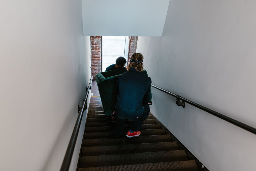 Two movers carry a green couch through a narrow stairwell, viewed from behind.
